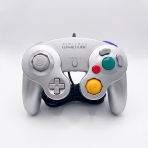 May include: A silver Nintendo GameCube video game controller featuring a central Start/Pause button, a gray analog stick, a directional pad, a large green A button, a red B button, a yellow C-stick, and gray X and Y buttons with a black cord.