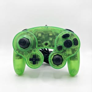 Puede incluir: Un mando de videojuegos de color verde neón translúcido con joysticks negros, una cruceta negra y botones de acción negros. Los circuitos internos son visibles a través de la carcasa de plástico transparente y tiene un cable negro conectado.