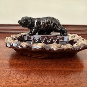 May include: A dark brown ceramic ashtray featuring a sculpted bear figure standing on the edge. The tray has a glossy finish with a wavy, textured rim that transitions into a lighter tan color, providing a contrast to the deep brown center.