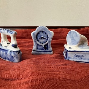 May include: Three miniature blue and white ceramic figurines: an iron, a clock, and a phonograph. The iron has a handle and decorative details. The clock features a round face. The phonograph has a horn-shaped speaker.