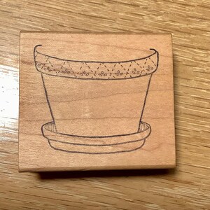 May include: A wooden stamp featuring a detailed illustration of a flower pot with a decorative rim and a matching saucer. The stamp is made of light-colored wood and is designed for crafting and scrapbooking projects.