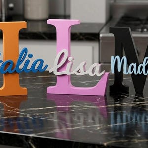 Op de afbeelding: Drie houten letters met namen. De oranje letter "I" heeft "Italia" in blauw schrift. De roze letter "L" heeft "Lisa" in wit schrift. De zwarte letter "M" heeft "Maddie" in lichtblauw schrift. Op een zwart marmeren oppervlak.