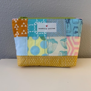May include: A quilted zipper pouch featuring a patchwork design of various fabrics in shades of mustard yellow, light blue, turquoise, and white. The front includes a white rectangular label with a small red heart and the text carefully quilted.