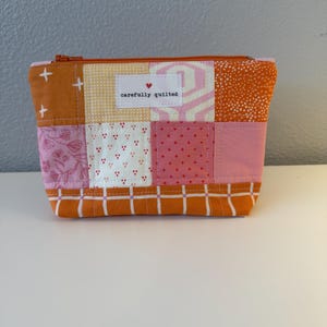 Peut inclure: Une pochette rectangulaire à fermeture éclair composée de pièces de tissu matelassé dans des tons orange, rose et blanc. L'avant présente une étiquette en tissu blanc avec un petit cœur rouge et le texte carefully quilted. Le bas est à carreaux orange.