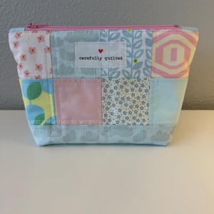 Könnte beinhalten: Ein Reißverschlusstäschchen aus gesteppten Stoffquadraten in Pastelltönen wie Rosa, Blau, Gelb und Weiß. Das Täschchen hat ein weißes, rechteckiges Etikett in der Mitte der Vorderseite mit einem kleinen roten Herz und dem Text carefully quilted.