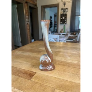 May include: A ceramic bud vase featuring a unique snail-like coiled base that transitions into a tall, slender neck. The piece is finished with a two-tone glaze, combining a warm terracotta brown at the bottom with a speckled cream white.