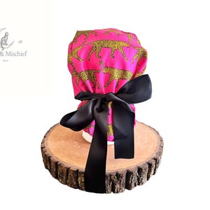 May include: A pink scrub cap with a gold leopard print, tied with a large black satin bow. The cap is displayed on a wooden slice. The logo "Moon & Mischief" is visible in the upper left corner.