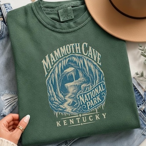 May include: A forest green crewneck sweatshirt featuring a graphic print of a cave interior with stalactites and the text Mammoth Cave National Park Kentucky. The shirt is displayed alongside blue denim jeans, a tan hat, and a gold necklace.