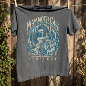 May include: A charcoal gray short-sleeve t-shirt featuring a blue and cream graphic of a cave interior. The text reads MAMMOTH CAVE NATIONAL PARK KENTUCKY. The design depicts stalactites and rock formations within a circular frame on the chest.