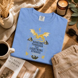 May include: A light blue t-shirt with a gold dragon graphic and the text "Dancing Through The Lightning Strikes". The shirt is folded on a wooden surface, next to a candle, sunglasses, and a magazine.