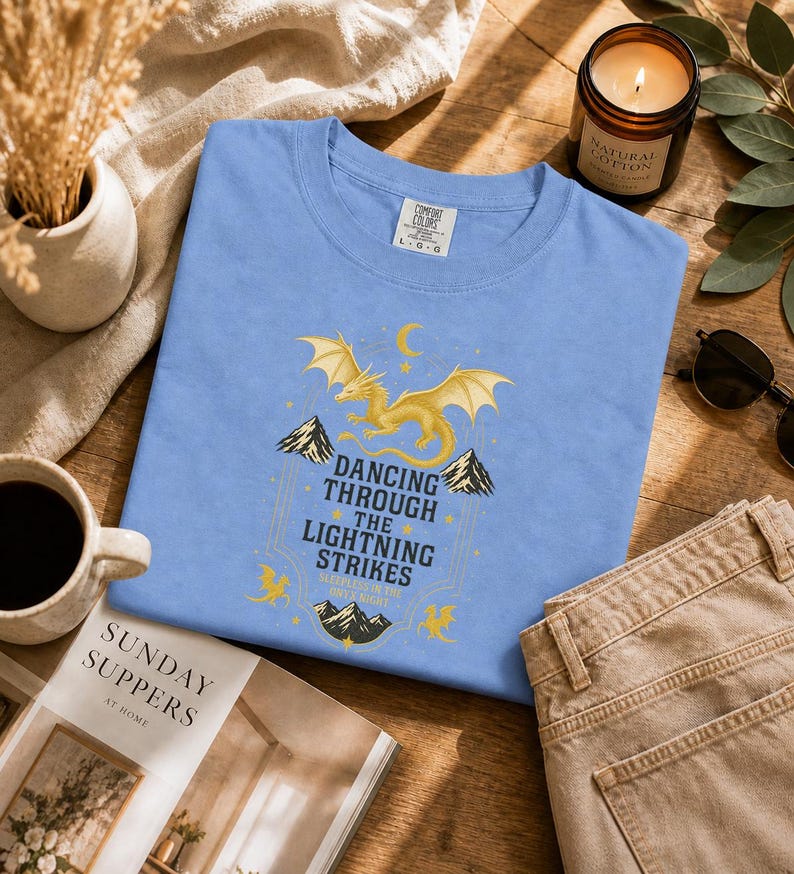 May include: A light blue t-shirt with a gold dragon graphic and the text "Dancing Through The Lightning Strikes". The shirt is folded on a wooden surface, next to a candle, sunglasses, and a magazine.