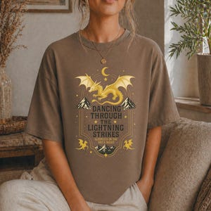 May include: Brown t-shirt featuring a golden dragon design with the text "Dancing Through The Lightning Strikes". The design includes mountains, stars, and a crescent moon. The shirt is a comfortable, casual wear.