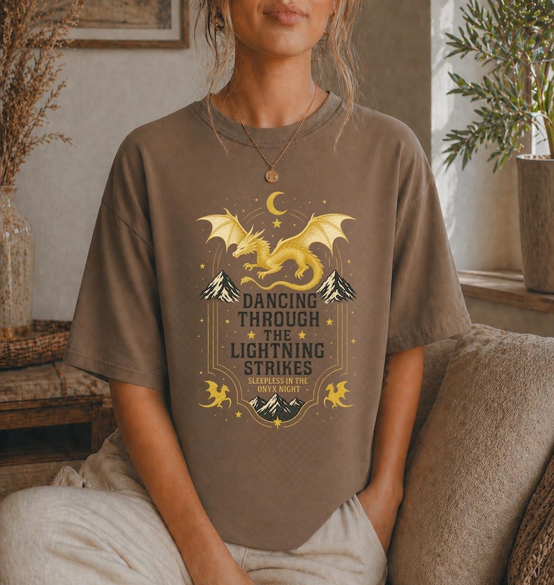 May include: Brown t-shirt featuring a golden dragon design with the text "Dancing Through The Lightning Strikes". The design includes mountains, stars, and a crescent moon. The shirt is a comfortable, casual wear.