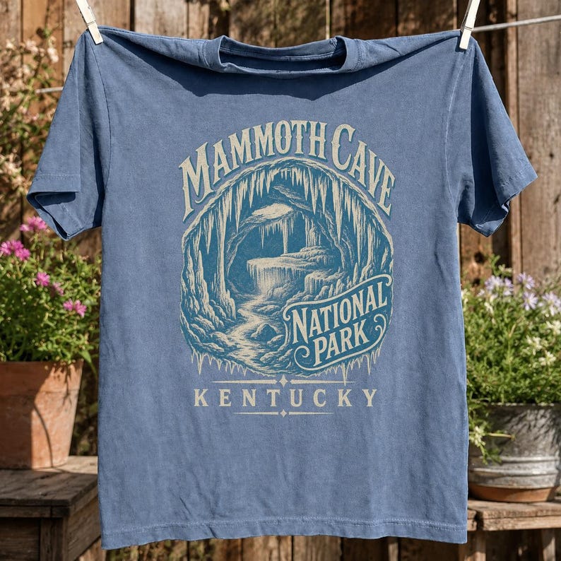 May include: A blue short-sleeve t-shirt featuring a graphic print of a cave interior with stalactites and stalagmites. The text reads MAMMOTH CAVE NATIONAL PARK KENTUCKY in a stylized font. The design is rendered in light blue and cream tones.