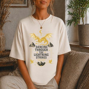 May include: Cream-colored t-shirt featuring a golden dragon design with the text "Dancing Through The Lightning Strikes". The graphic includes mountains, stars, and a crescent moon. The shirt has short sleeves and a relaxed fit.