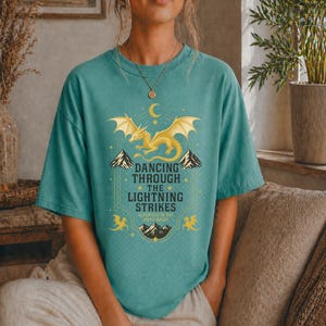 May include: Teal-colored t-shirt featuring a golden dragon design with the text "Dancing Through The Lightning Strikes." The design includes mountains, stars, and a crescent moon. The shirt is a comfortable, casual wear item.