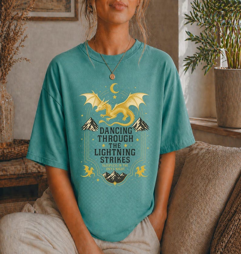 May include: Teal-colored t-shirt featuring a golden dragon design with the text "Dancing Through The Lightning Strikes." The design includes mountains, stars, and a crescent moon. The shirt is a comfortable, casual wear item.