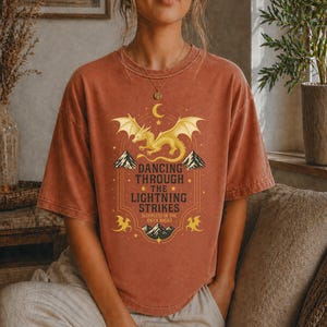 May include: Rust-colored t-shirt featuring a golden dragon design with the text "Dancing Through The Lightning Strikes". The graphic includes mountains, stars, and a crescent moon. The shirt is a short-sleeved, crew neck style.