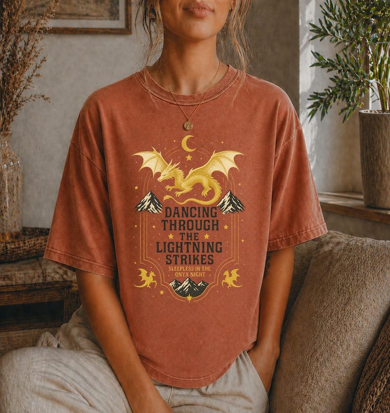 May include: Rust-colored t-shirt featuring a golden dragon design with the text "Dancing Through The Lightning Strikes". The graphic includes mountains, stars, and a crescent moon. The shirt is a short-sleeved, crew neck style.