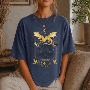 May include: Navy blue t-shirt featuring a golden dragon design with the text "Dancing Through The Lightning Strikes". The design includes mountains, stars, and a crescent moon. The shirt is a relaxed fit.