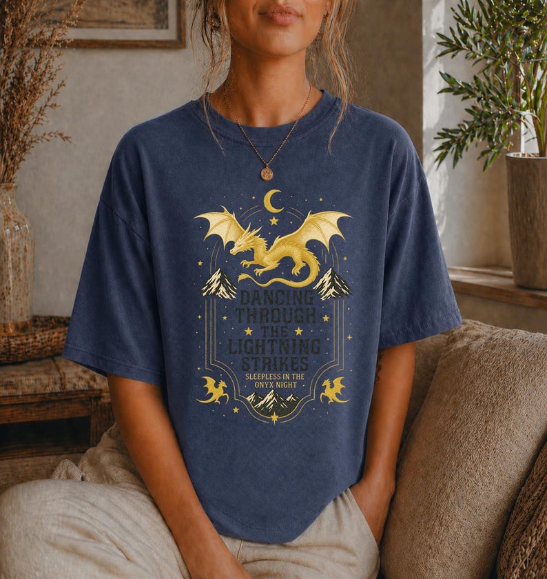 May include: Navy blue t-shirt featuring a golden dragon design with the text "Dancing Through The Lightning Strikes". The design includes mountains, stars, and a crescent moon. The shirt is a relaxed fit.