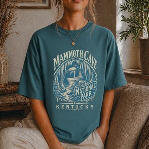 May include: A teal short-sleeve t-shirt featuring a cream-colored graphic of a cave interior. The text reads MAMMOTH CAVE NATIONAL PARK KENTUCKY. The design depicts stalactites and rock formations within a circular frame on the front of the shirt.