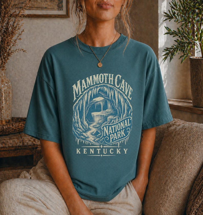 May include: A teal short-sleeve t-shirt featuring a cream-colored graphic of a cave interior. The text reads MAMMOTH CAVE NATIONAL PARK KENTUCKY. The design depicts stalactites and rock formations within a circular frame on the front of the shirt.