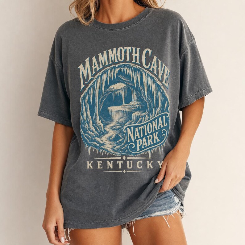 May include: A charcoal gray short-sleeve t-shirt featuring a blue graphic print of a cave interior with stalactites. The text reads MAMMOTH CAVE NATIONAL PARK KENTUCKY in a vintage-style font. The shirt has a crew neckline and a relaxed fit.