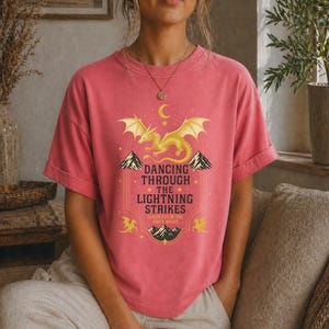 May include: A coral-colored t-shirt featuring a golden dragon design with the text "Dancing Through The Lightning Strikes" and "Sleepless in the Onyx Night". The design includes mountains, stars, and a crescent moon.