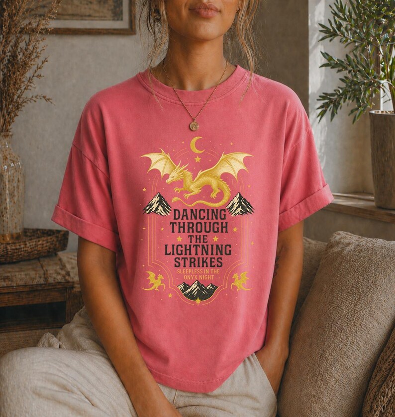 May include: A coral-colored t-shirt featuring a golden dragon design with the text "Dancing Through The Lightning Strikes" and "Sleepless in the Onyx Night". The design includes mountains, stars, and a crescent moon.