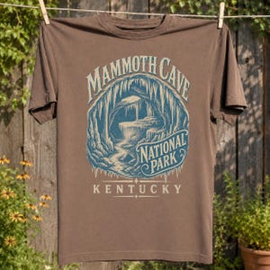 May include: A brown short-sleeve t-shirt featuring a blue and cream graphic of a cave interior. The text reads MAMMOTH CAVE NATIONAL PARK KENTUCKY. The design depicts stalactites and rock formations within a circular frame on the front of the shirt.