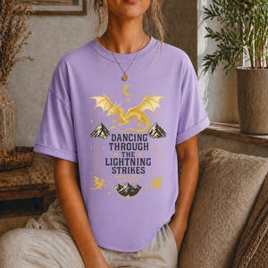 May include: A lilac-colored t-shirt featuring a golden dragon design with the text "Dancing Through The Lightning Strikes". The graphic includes mountains, stars, and a crescent moon. The shirt is a comfortable, casual style.
