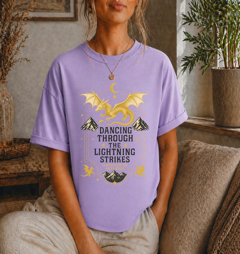 May include: A lilac-colored t-shirt featuring a golden dragon design with the text "Dancing Through The Lightning Strikes". The graphic includes mountains, stars, and a crescent moon. The shirt is a comfortable, casual style.