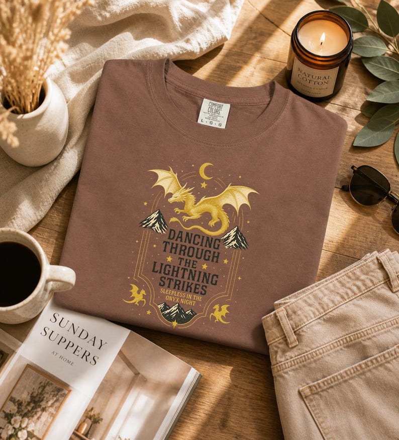 May include: A brown t-shirt with a golden dragon graphic and the text "Dancing Through The Lightning Strikes." The shirt is displayed on a wooden surface with a lit candle, sunglasses, and a magazine.