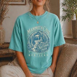 May include: A teal short-sleeve t-shirt featuring a graphic of a cave interior with stalactites and a stream. The text reads MAMMOTH CAVE NATIONAL PARK KENTUCKY in a vintage-style font. The design is printed in shades of cream and dark blue.