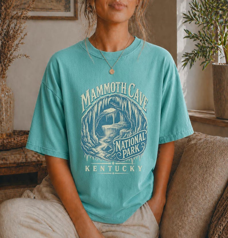 May include: A teal short-sleeve t-shirt featuring a graphic of a cave interior with stalactites and a stream. The text reads MAMMOTH CAVE NATIONAL PARK KENTUCKY in a vintage-style font. The design is printed in shades of cream and dark blue.