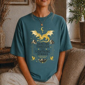 May include: Teal-colored t-shirt featuring a golden dragon design with the text "Dancing Through The Lightning Strikes". The graphic includes a crescent moon, mountains, and stars. The shirt is a casual, oversized fit.