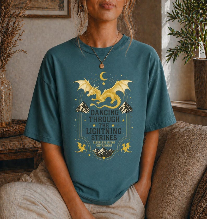 May include: Teal-colored t-shirt featuring a golden dragon design with the text "Dancing Through The Lightning Strikes". The graphic includes a crescent moon, mountains, and stars. The shirt is a casual, oversized fit.