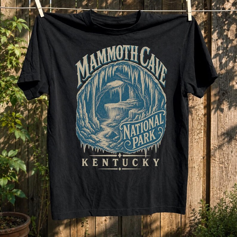 May include: A black short-sleeve t-shirt featuring a blue and cream graphic of a cave interior with stalactites. The text reads MAMMOTH CAVE NATIONAL PARK KENTUCKY in a stylized font. The design is centered on the chest of the garment.
