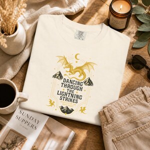 May include: Cream-colored t-shirt with a gold dragon graphic and the text "Dancing Through The Lightning Strikes". The shirt is made of a soft material and is folded on a wooden surface.