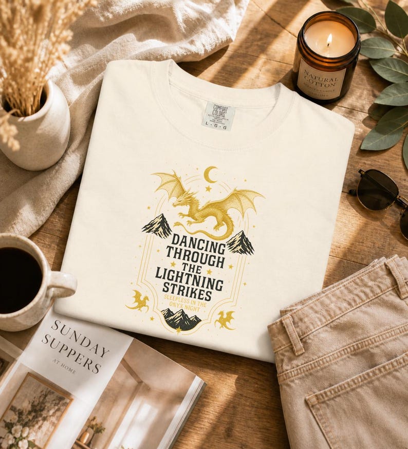 May include: Cream-colored t-shirt with a gold dragon graphic and the text "Dancing Through The Lightning Strikes". The shirt is made of a soft material and is folded on a wooden surface.