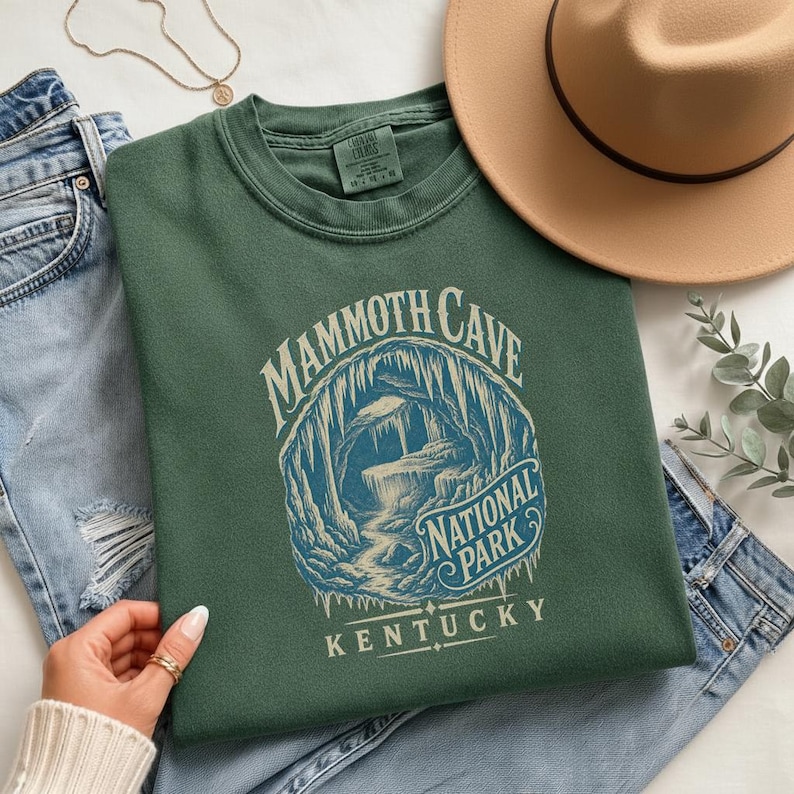 May include: A forest green crewneck sweatshirt featuring a graphic print of a cave interior with stalactites and the text Mammoth Cave National Park Kentucky. The shirt is displayed alongside blue denim jeans, a tan hat, and a gold necklace.