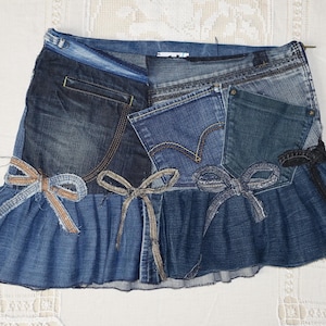 Peut inclure: Jupe en jean patchwork avec un design superposé. La jupe présente différentes nuances de denim bleu, des poches et des nœuds décoratifs. La jupe a un ourlet à volants et est présentée sur une surface blanche.