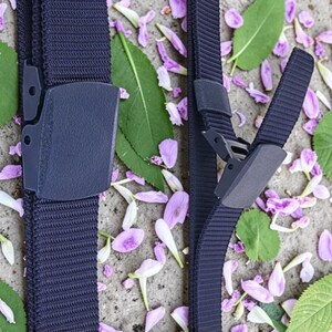 May include: A dark navy blue nylon webbing belt featuring a matching plastic buckle. The belt strap has a textured, ribbed surface, and the buckle mechanism is shown in both a closed, locked position and an open, unfastened state.