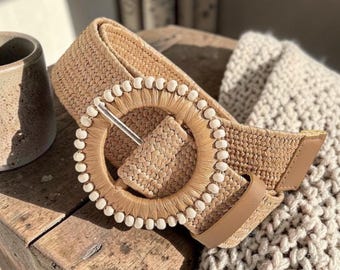 May include: A woven beige belt with a large circular buckle. The buckle is adorned with small white beads around the outer edge. The belt has a tan leather tab and is resting on a wooden surface.
