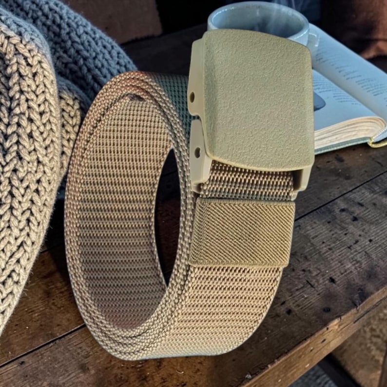 May include: A tan webbed belt featuring a matching rectangular plastic buckle. The belt is constructed from a durable woven fabric with a textured surface and includes a small elastic loop to secure the excess strap once fastened around the waist.