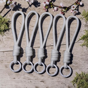 Può includere: Cinque cordini in paracord bianchi, ciascuno caratterizzato da un'asola fissata con un nodo strettamente avvolto e collegata a un anello circolare in metallo bianco con chiusura a molla. I cordini sono disposti in fila in modo uniforme.