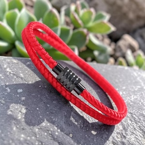 Könnte beinhalten: Ein leuchtend rotes geflochtenes Armband mit einem schwarzen Magnetverschluss. Das Armband aus dickem, gewebtem Material wird auf einer grauen Steinoberfläche mit grünem Laub im Hintergrund präsentiert. Ein modisches Accessoire.