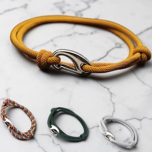 Könnte beinhalten: Eine Kollektion von vier Armbändern aus Kordelmaterial, die mit einem silberfarbenen Metallkarabiner verschlossen werden. Ein Armband ist senfgelb, eines ist mehrfarbig mit Rot, Gelb und Grün, eines ist dunkelgrün und eines ist weiß.