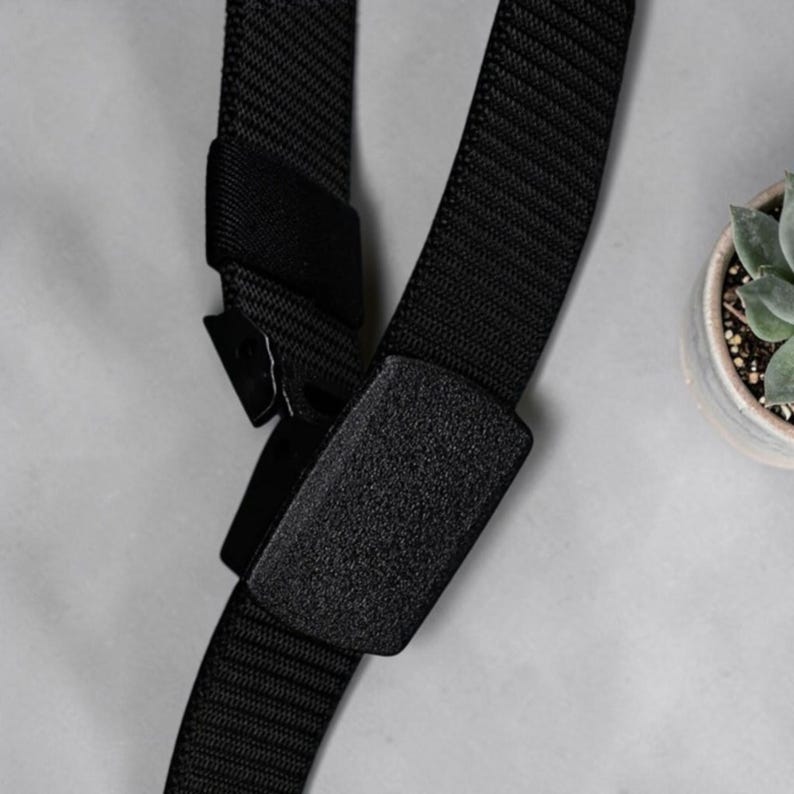 May include: A black nylon webbing belt featuring a rectangular plastic buckle with a textured matte finish. The belt strap is constructed from durable woven fabric and includes a matching black plastic sliding mechanism for secure fastening.